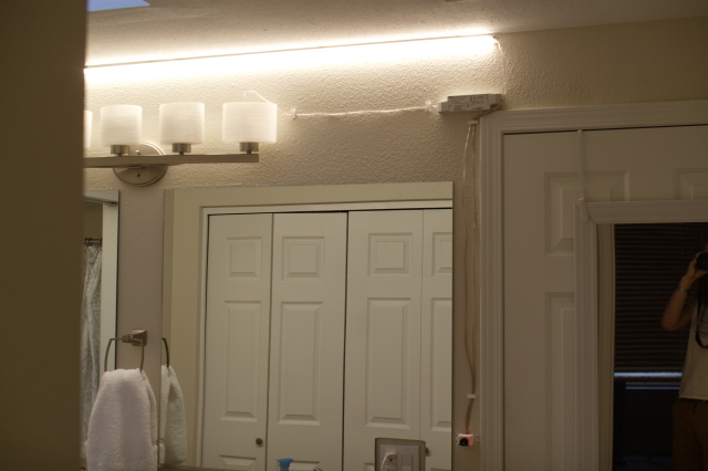 Bathroom lights, above old light fixture and mirror, with photographer in a mirror dimly off to the side